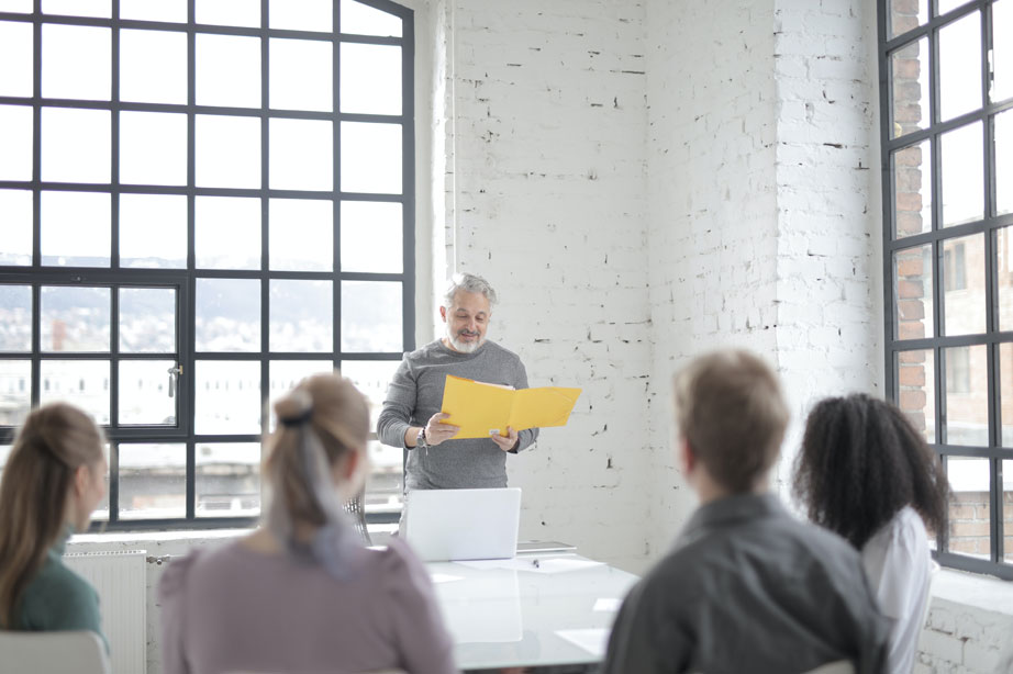 Ein älterer Mann steht mit einem gelben Ordner am Kopfende eines Tisches und spricht während einer Betriebsratssitzung zu drei sitzenden Kollegen in einem hellen, modernen Büro mit großen Fenstern und weißen Backsteinwänden. - RUF Beratung - Ihre Contact Center Berater
