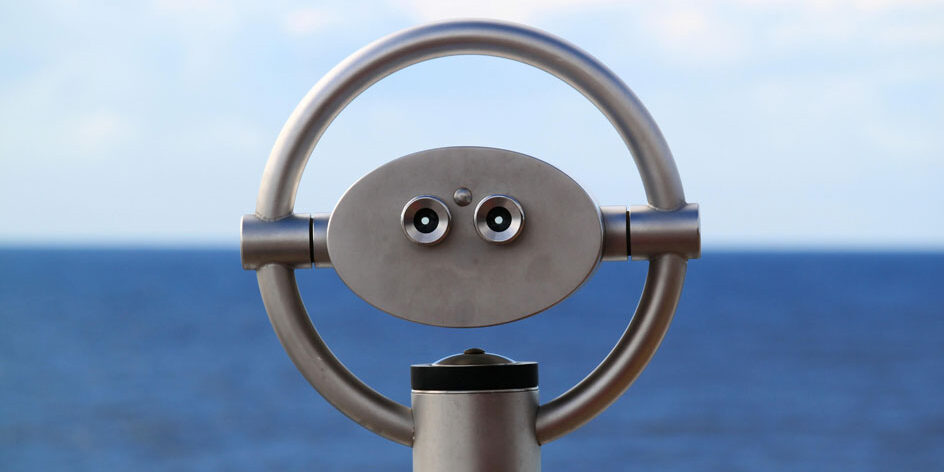 Ein silberner, münzbetriebener Binokular-Viewer, der einem Gesicht mit zwei Augen ähnelt, steht einsatzbereit – ähnlich wie Externe Audits Klarheit bieten – vor dem verschwommenen blauen Ozean und Himmel. - RUF Beratung - Ihre Contact Center Berater