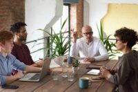 Vier Personen sitzen in einem modernen Büro um einen Holztisch und diskutieren über Personalmanagement. Laptops, Notizbücher, Tassen und Pflanzen stehen auf dem Tisch. Eine Person gestikuliert beim Sprechen; die anderen hören aufmerksam zu. - RUF Beratung - Ihre Contact Center Berater