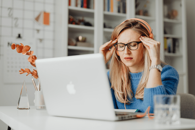Eine Frau mit Brille und Kopfhörern sitzt mit einem Laptop an einem Schreibtisch, hält sich die Schläfen und wirkt gestresst, möglicherweise aufgrund der Anforderungen des Intraday-Managements. Auf dem Schreibtisch stehen Büromaterialien und ein Glas Wasser. - RUF Beratung - Ihre Contact Center Berater