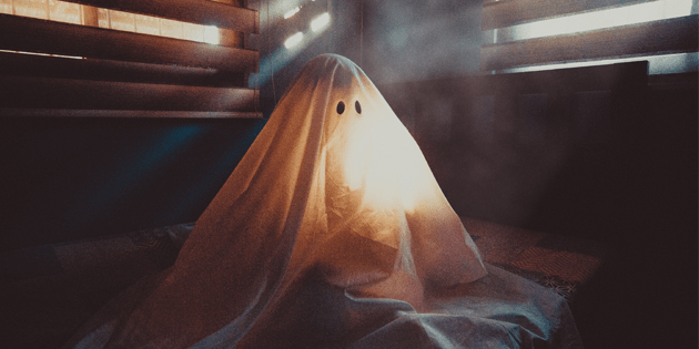 Eine Person, die mit einem weißen Laken mit zwei schwarzen Augenlöchern bedeckt ist und einem Geist ähnelt, sitzt auf einem Bett in einem schwach beleuchteten Raum mit Jalousien, die Lichtstreifen hereinlassen und so die stille Einsamkeit hervorrufen, die oft in CX-Kontaktcenter-Umgebungen herrscht. - RUF Beratung - Ihre Contact Center Berater