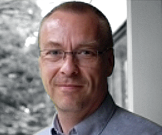 A man with short hair and glasses, wearing a collared shirt, smiles slightly while standing indoors near a window with blurred trees visible outside. - RUF Beratung - Ihre Contact Center Berater