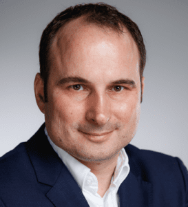 A man with short dark hair, wearing a navy blue suit jacket and a white shirt, smiles slightly while looking at the camera against a plain gray background. - RUF Beratung - Ihre Contact Center Berater