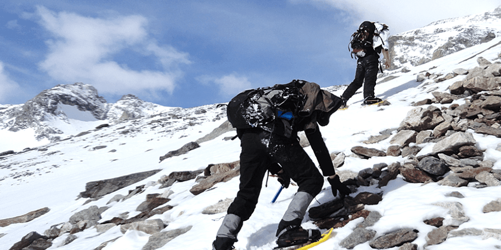 Zwei Bergsteiger in Winterkleidung und mit Rucksäcken erklimmen unter einem teilweise bewölkten Himmel einen schneebedeckten, felsigen Berghang, umgeben von schneebedeckten Gipfeln. - RUF Beratung - Ihre Contact Center Berater