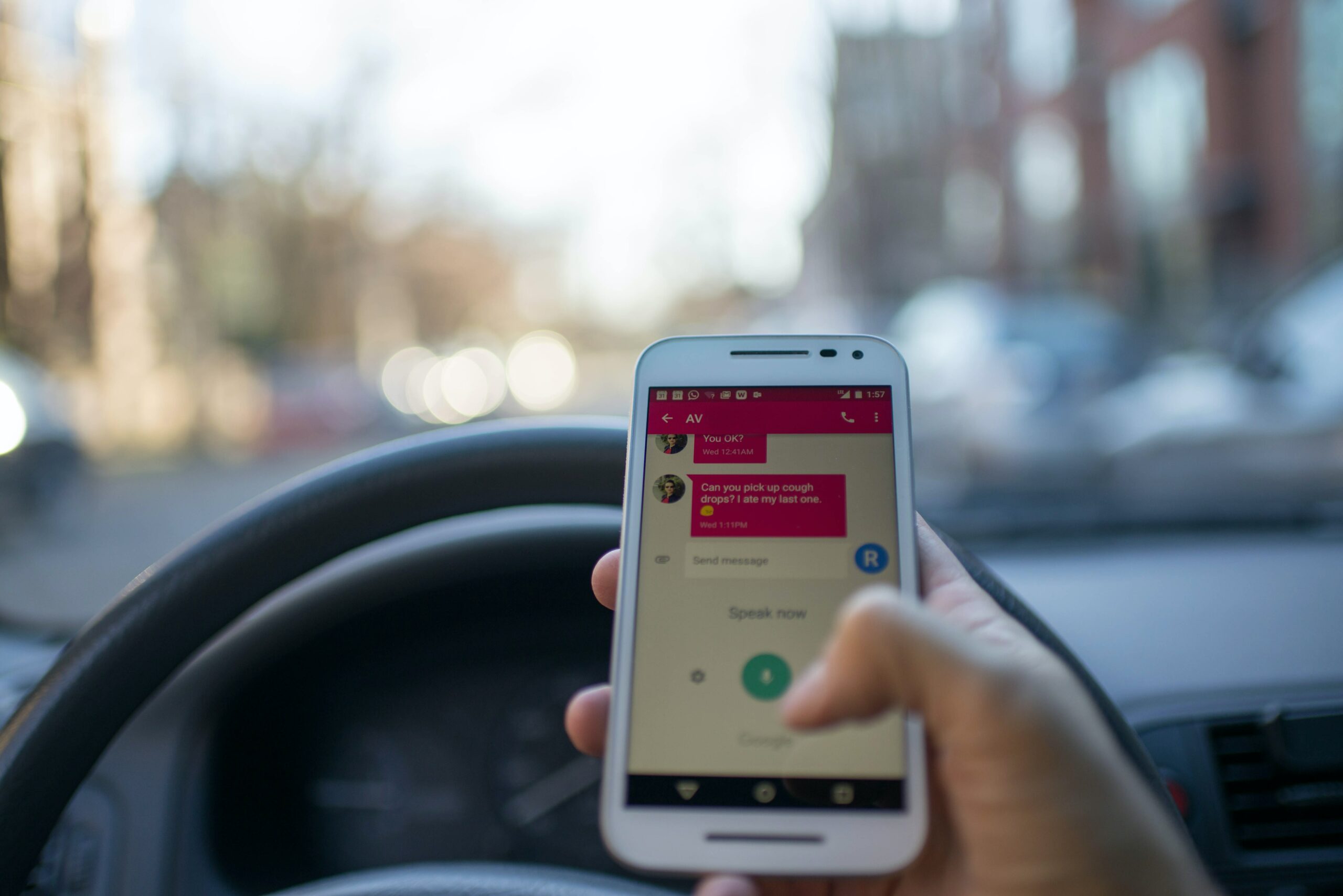 Eine Person hält im Auto ein Smartphone und tippt mit einer Hand eine SMS, während sie hinter dem Lenkrad sitzt. Auf dem Telefonbildschirm wird ein Chat über Automatisierung für Kunden angezeigt; die Außenansicht ist verschwommen. - RUF Beratung - Ihre Contact Center Berater