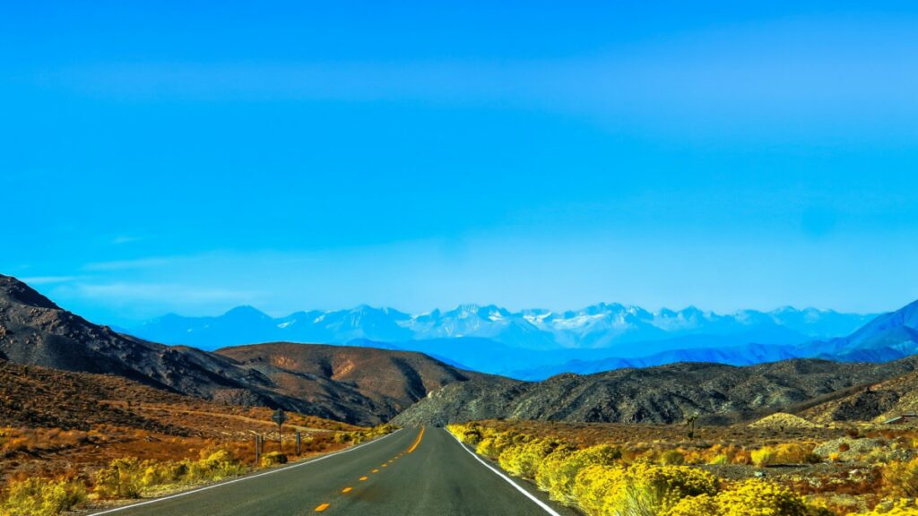 Eine gerade, leere Straße erstreckt sich durch trockene, felsige Hügel mit gelber Vegetation und führt zu schneebedeckten Bergen in der Ferne unter einem strahlend blauen Himmel – ein Bild, das die Customer Journey widerspiegelt. Eine Einführung. - RUF Beratung - Ihre Contact Center Berater