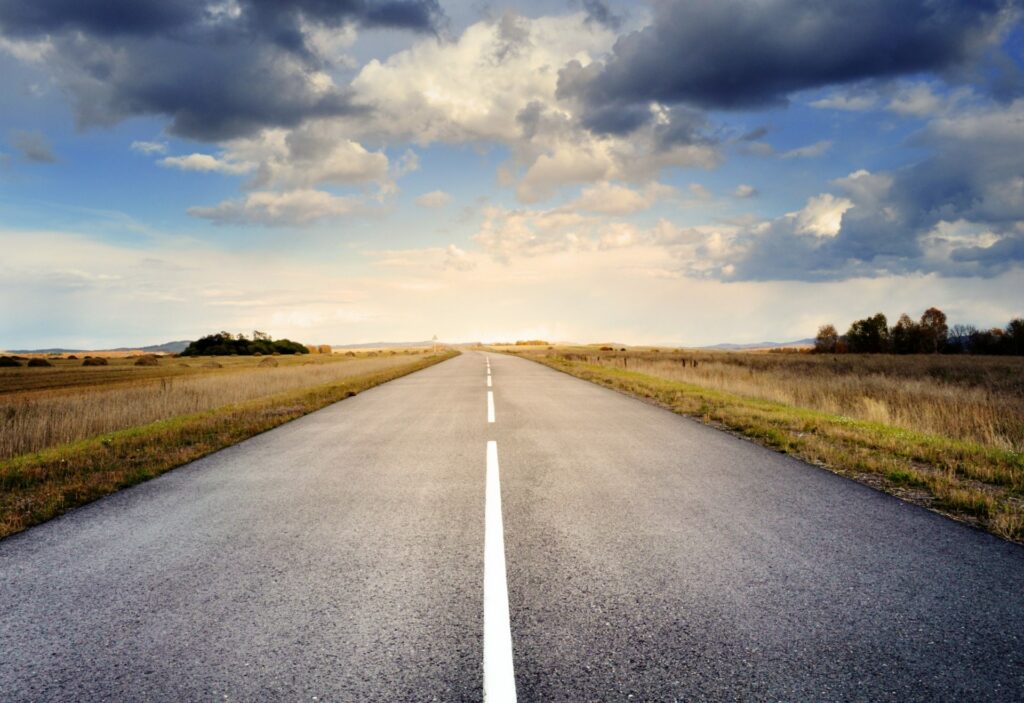 Eine gerade, leere Straße erstreckt sich in der Ferne durch flache Felder unter einem teilweise bewölkten Himmel, durch den das Sonnenlicht bricht – die perfekte visuelle Metapher für eine klare Customer Journey-Spezifikation, die sich vor uns entfaltet. - RUF Beratung - Ihre Contact Center Berater