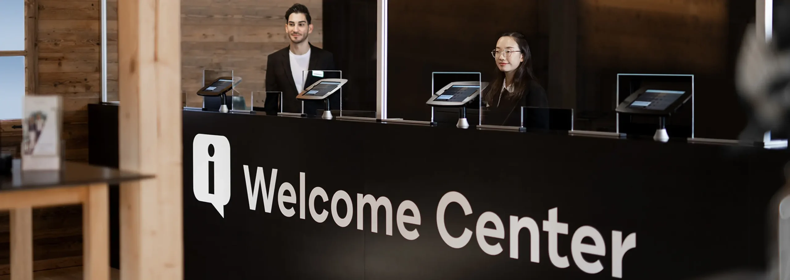 Zwei Rezeptionisten, ein Mann und eine Frau, stehen hinter einem schwarzen Tresen mit der Aufschrift „Welcome Center“ und Tablets zur Dienstleister-Steuerung in einer modernen Lobby mit Holzakzenten. - RUF Beratung - Ihre Contact Center Berater