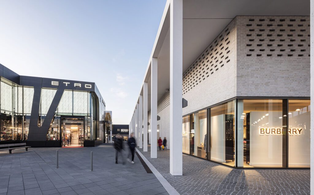 Modernes Outdoor-Einkaufszentrum mit großen Glasfassaden für Etro und Burberry. Die Menschen flanieren auf breiten, von hohen weißen Säulen und Backsteinmauern gesäumten Wegen unter klarem Himmel, während Dienstleister-Steuerung für ein reibungsloses Facility Management sorgt. - RUF Beratung - Ihre Contact Center Berater