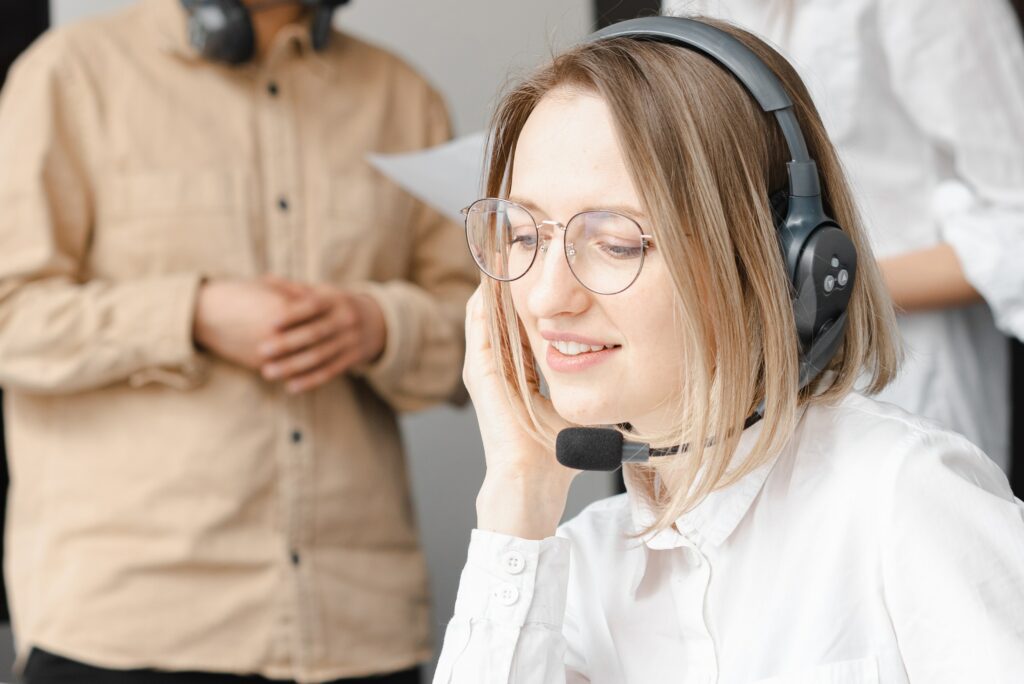 Eine Frau mit Brille und Headset lächelt an einem Schreibtisch und demonstriert so ihre erfolgreiche Mitarbeiterbindung im Kundenservice. Im Hintergrund steht eine weitere Person, ebenfalls mit Headset. Beide wirken in ihrer Büroumgebung engagiert. - RUF Beratung - Ihre Contact Center Berater