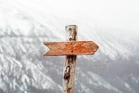 Ein verwitterter Holzwegweiser mit einem roten Pfeil zeigt nach rechts, vor einem verschwommenen, schneebedeckten Berghintergrund – und lädt Sie zu einem Kundenservice-Webinar zum EU AI Act ein. - RUF Beratung - Ihre Contact Center Berater