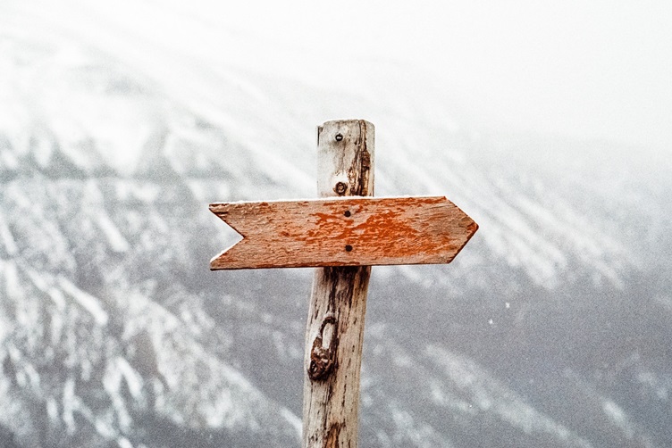 Ein verwitterter Holzwegweiser mit einem roten Pfeil zeigt nach rechts, vor einem verschwommenen, schneebedeckten Berghintergrund – und lädt Sie zu einem Kundenservice-Webinar zum EU AI Act ein. - RUF Beratung - Ihre Contact Center Berater