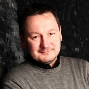 A man with short dark hair and slight stubble smiles gently at the camera. He is wearing a grey sweater over a black shirt and stands against a dark, textured background, reflecting the approachable style of RUF Beratung. - RUF Beratung - Ihre Contact Center Berater