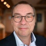Ein Bild von Arno Rebmann mit kurzen braunen Haaren, Brille, weißem Hemd und dunklem Blazer lächelt in einem sanft beleuchteten Innenbereich warm in die Kamera – und spiegelt damit die zugängliche Professionalität eines Contact Center-Beraters wider. - RUF Beratung - Ihre Contact Center Berater