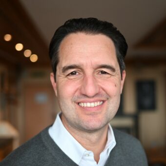 A man with short dark hair and a gray sweater over a white collared shirt smiles at the camera in a warmly lit indoor setting, reflecting the approachable professionalism of RUF Beratung. - RUF Beratung - Ihre Contact Center Berater