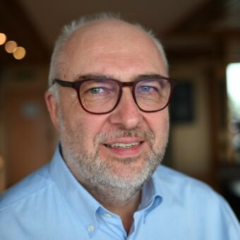 Ein Bild von Peter Basse mit Brille, kurzen grauen Haaren und Bart sowie einem hellblauen Hemd steht in einem warm beleuchteten Innenbereich – ein selbstbewusster Contact Center-Berater vor einem leicht verschwommenen Hintergrund. - RUF Beratung - Ihre Contact Center Berater