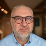 Ein Bild von Peter Basse mit Brille, kurzen grauen Haaren und Bart sowie einem hellblauen Hemd steht in einem warm beleuchteten Innenbereich – ein selbstbewusster Contact Center-Berater vor einem leicht verschwommenen Hintergrund. - RUF Beratung - Ihre Contact Center Berater