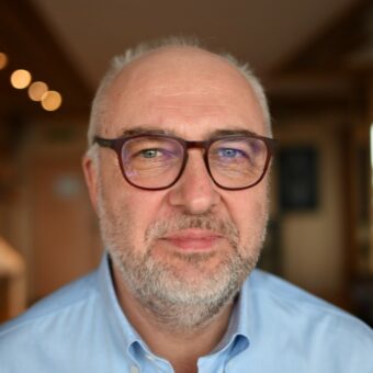 A middle-aged man with glasses and a gray beard, representing RUF Beratung, is wearing a light blue collared shirt. He is looking at the camera and smiling softly against a warmly lit, softly blurred background. - RUF Beratung - Ihre Contact Center Berater
