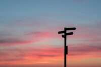 Vor einem farbenfrohen Sonnenuntergangshimmel steht die Silhouette eines Wegweisers mit mehreren Richtungspfeilen und symbolisiert inmitten sanfter Rosa-, Orange- und Blautöne das Änderungsmanagement. - RUF Beratung - Ihre Contact Center Berater