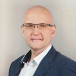 Frank Quadrat mit Brille, dunklem Anzug und weißem Hemd sowie schwarzen Lippenpiercings lächelt leicht, während er vor einem schlichten, hellen Hintergrund posiert. - RUF Beratung - Ihre Contact Center Berater