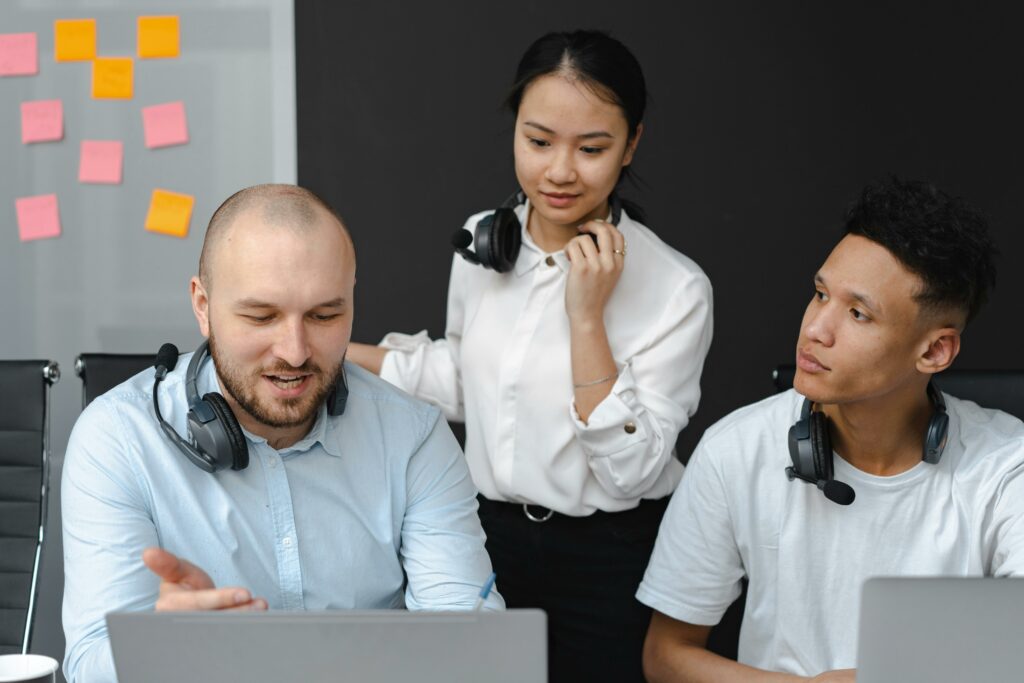Drei Personen arbeiten mit Laptops und Headsets an einem Schreibtisch und konzentrieren sich auf die Mitarbeiterqualifizierung. Ein Mann gestikuliert beim Sprechen, eine Frau steht daneben und hört zu, ein weiterer Mann schaut aufmerksam zu. An der Wand hinter ihnen hängen bunte Haftnotizen. - RUF Beratung - Ihre Contact Center Berater