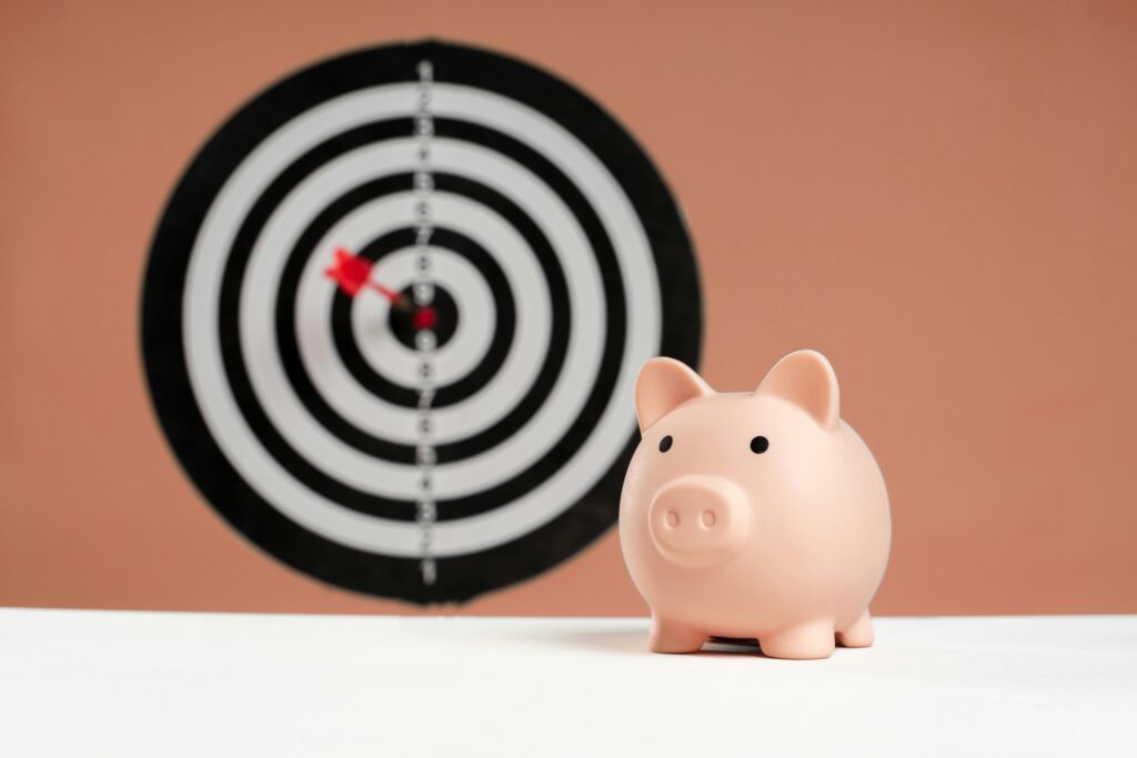 Im Vordergrund steht auf einer weißen Fläche ein Sparschwein, im Hintergrund eine schwarz-weiße Dartscheibe – mit einem einzelnen roten Pfeil in der Mitte –, die vor einer pfirsichfarbenen Wand Präzision während der Customer Experience Week symbolisiert. - RUF Beratung - Ihre Contact Center Berater