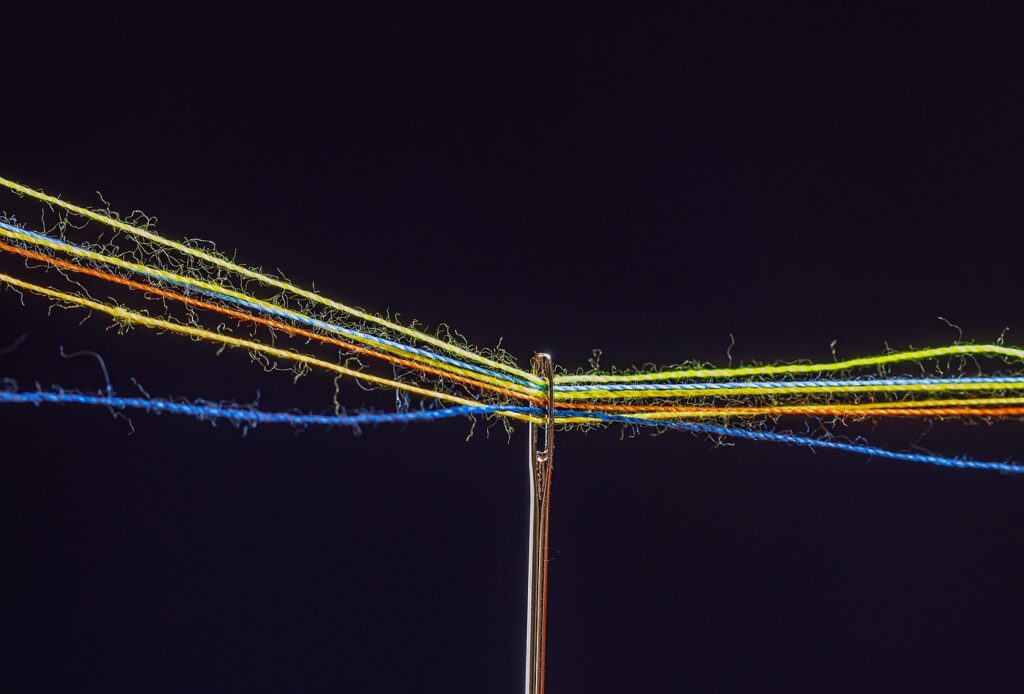 Eine Nahaufnahme einer Nadel, deren Öhr mit mehreren bunten Fäden durchzogen ist, darunter Gelb, Blau, Orange und Grün, vor einem dunklen Hintergrund – ein Symbol dafür, wie Omnichannel die Stränge des modernen Kundenservice verbindet. - RUF Beratung - Ihre Contact Center Berater