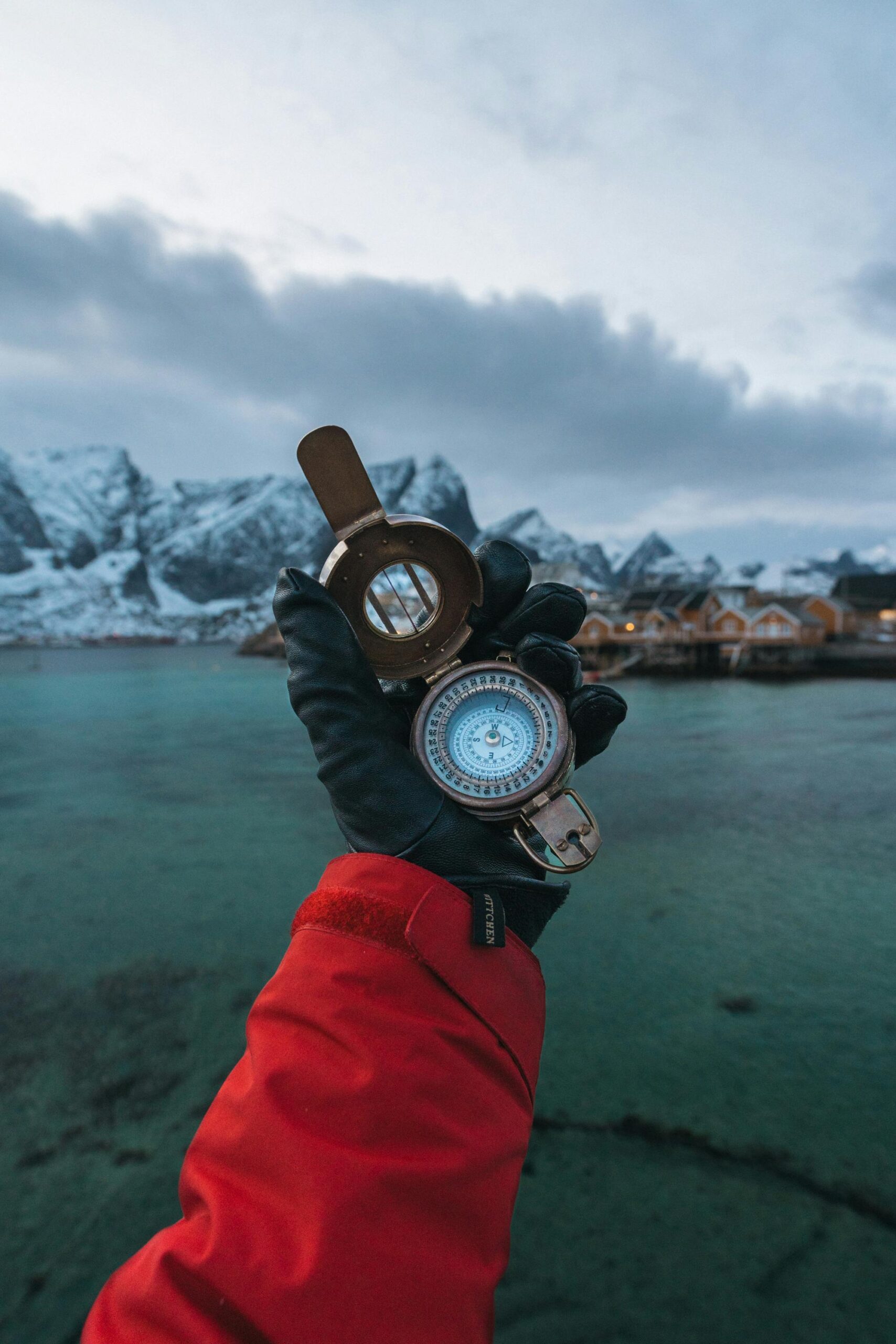 Eine behandschuhte Hand in einer roten Jacke hält einen offenen Kompass vor schneebedeckten Bergen, türkisfarbenem Wasser und Hütten unter einem bewölkten Himmel – und fängt den Entdeckergeist ein, der den CX Dialog 2025 inspiriert. - RUF Beratung - Ihre Contact Center Berater