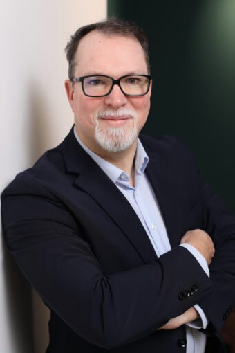 A middle-aged man with glasses, a short gray beard, and mustache, wearing a navy blazer and light blue shirt, stands with arms crossed and smiles while leaning against a white wall. - RUF Beratung - Ihre Contact Center Berater