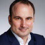 A man with short dark hair, wearing a navy blue suit jacket and a white shirt, smiles slightly while looking at the camera against a plain gray background. - RUF Beratung - Ihre Contact Center Berater